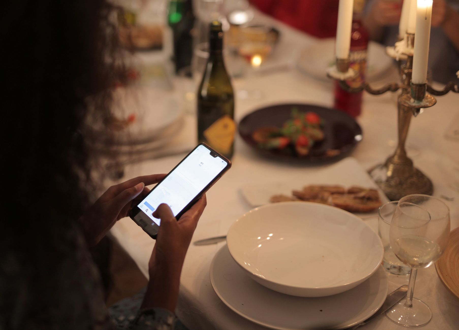 A cozy dinner setting with a woman using a smartphone at a candlelit table.